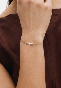 Gouden armband met het woord "LOVE" in blokletters, verbonden door een fijn kettinkje, gedragen om een pols tegen een bruine achtergrond.