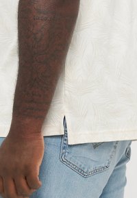 Maglietta a maniche corte in crema con un delicato motivo di foglie di palma, abbinata a jeans in denim azzurro chiaro. Presenta fessure laterali per maggiore comfort.