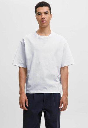 Mann, der nach vorne steht und ein einfaches, übergroßes weißes T-Shirt sowie lockere, dunkle Hosen trägt, vor einem schlichten, hellen Hintergrund.