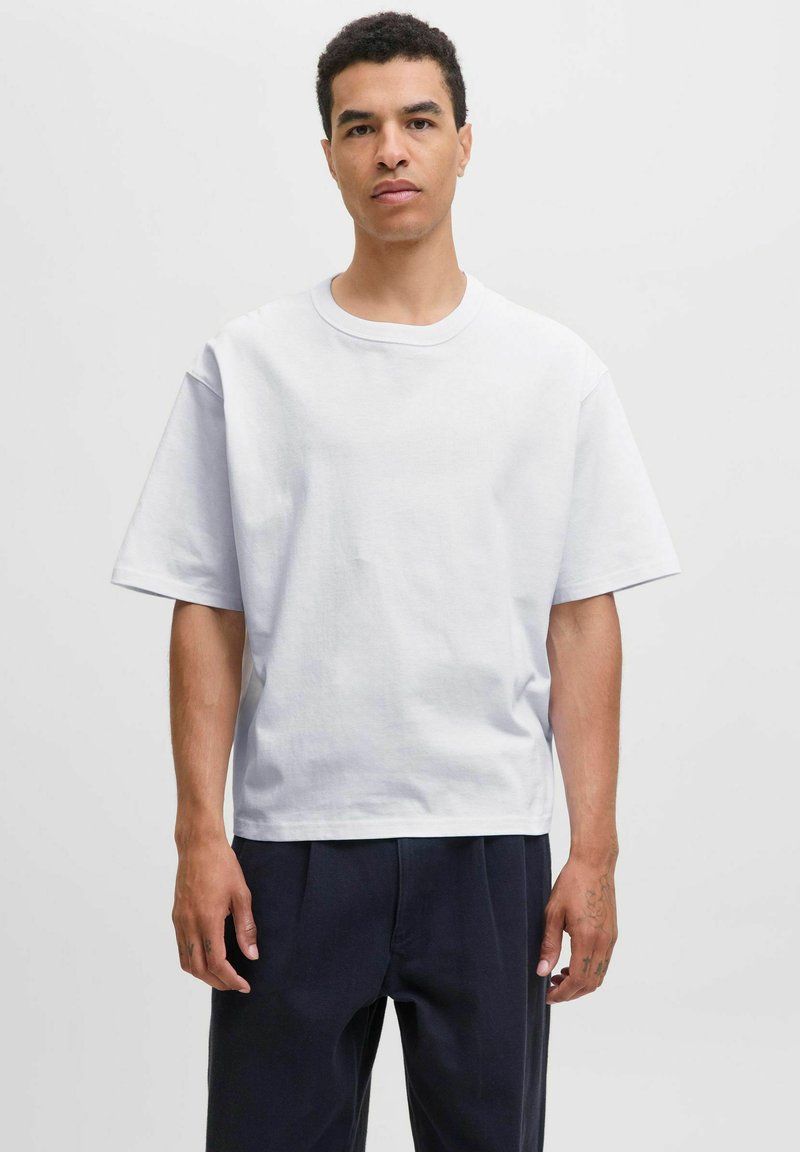 Bărbat în picioare, cu fața la cameră, îmbrăcat cu un tricou alb simplu, supradimensionat, și pantaloni clari, largi, pe un fundal simplu, deschis la culoare.