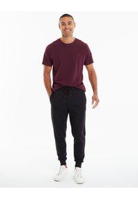 Burgunderfarbenes Kurzarm-T-Shirt kombiniert mit schwarzen Jogginghosen. Die Jogginghosen verfügen über einen Kordelzug und gerippte Bündchen. Weiße Sneakers runden den Look ab.