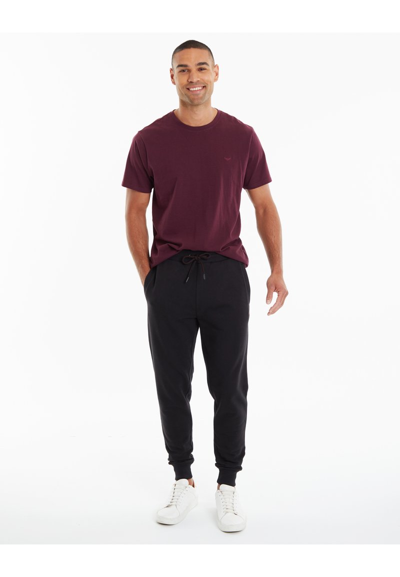 Burgunderfarbenes Kurzarm-T-Shirt kombiniert mit schwarzen Jogginghosen. Die Jogginghosen verfügen über einen Kordelzug und gerippte Bündchen. Weiße Sneakers runden den Look ab.
