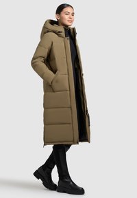 Lunga giacca invernale verde oliva con cappuccio, caratterizzata da un design trapuntato, tasche laterali e chiusura con zip. Abbinata a stivali neri fino al ginocchio.