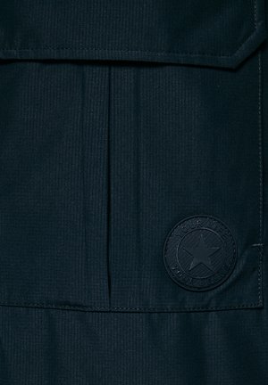 Tamno plava tkanina s blagim reljefnim uzorkom; sadrži prošiveni džep s preklopom i okruglu utisnutu oznaku s tekstom i dizajnom zvijezde.