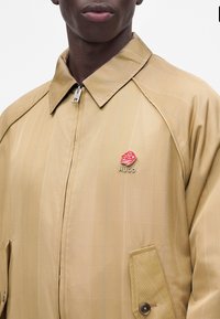 Gros plan d’une personne portant une veste beige zippée avec col et une épingle rose rouge portant la mention "HUGO" sur la poitrine.