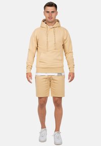 Felpa beige con tasca frontale e lacci, abbinata a pantaloni corti beige coordinati. Entrambi realizzati in tessuto morbido, indossati con sneaker bianche.