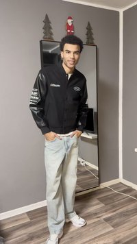 Μαύρο μπουφάν bomber με δερμάτινα μανίκια, διακοσμημένο με λευκά λογότυπα. Συνδυασμένο με ανοιχτό μπλε τζιν και λευκά αθλητικά παπούτσια, όρθιος σε ένα δωμάτιο.