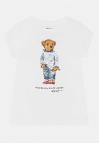 Bílé tričko s krátkým rukávem s grafikou medvěda ve modrém svetru, džínech a červených teniskách, s textem "Polo Bear by Ralph Lauren".