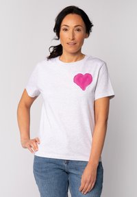 T-shirt blanc à manches courtes avec un grand motif de cœur rose sur la poitrine gauche, associé à un jean en denim bleu.