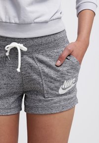 Šedé Nike šortky vyrobené z měkké, texturaé látky. Vlastnosti zahrnují bílé logo, pas na šňůrku a boční kapsy pro volnočasové nošení.