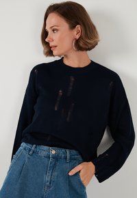 Tengeri kék kötött pulóver bordás textúrával és kopott részletekkel, magasan szabott kék farmernadrággal párosítva. Egyszerű, kerek nyakkivágás és laza szabás.