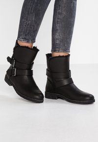 Botas de tobillo negras de cuero con un suave forro de piel. Cuentan con dos correas ajustables y una suela de goma texturizada.