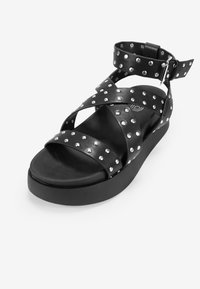 Black leather sandal with stud accents, cross straps, padded sole, and an adjustable ankle strap. Thick platform base for support.