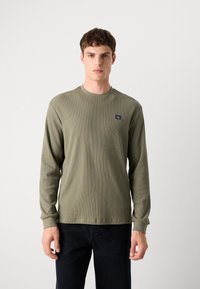 Olijfgroene ribbed shirt met lange mouwen en ronde halslijn, voorzien van een geborduurd logo op de borst. Zachte textuur en aansluitend ontwerp.