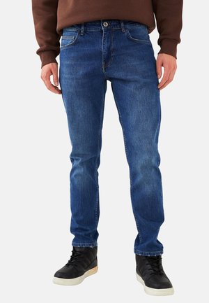 Mann trägt blaue Slim-Fit-Jeans, schwarze High-Top-Sneaker und einen braunen Pullover, steht mit locker an den Seiten hängenden Händen.