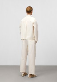 Giacca di cotone color beige chiaro con una vestibilità rilassata e piega sul retro, abbinata a pantaloni larghi coordinati, dotati di due tasche posteriori.