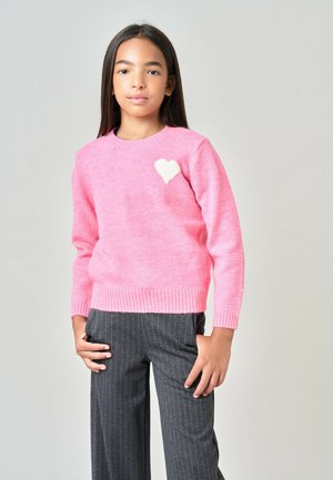 Meisje met steil donker haar dat een roze trui met een wit hart en grijze fijngestreepte broek draagt tegen een neutrale achtergrond.