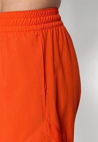 Oranžinės, lengvos sportinės šortai su elastine juosta, turintys šoninę kišenę ir geometrinio siuvinėjimo detales ant kojos.