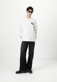 Hvid sweatshirt med grafisk print, mørke vide jeans og sorte sneakers. Modellen bærer runde, mørke solbriller og står foran en ensfarvet baggrund.