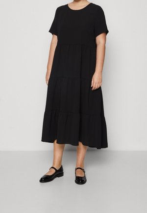 Femme portant une robe midi noire à manches courtes avec jupe à volants et chaussures Mary Jane noires, debout devant un fond clair uni.