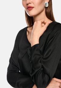 Frau, die eine schwarze Satinbluse mit langen Ärmeln trägt, hält eine Hand in der Nähe ihres Schlüsselbeins, trägt silberne quadratische Ohrringe und roten Lippenstift.