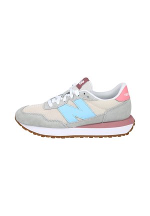 Zapatilla gris y beige con logo "N" en azul claro, cordones blancos, lengüeta rosa en el talón y suela marrón acanalada, fotografiada desde el lateral.