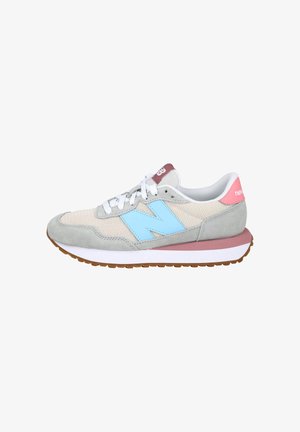 Zapatilla gris y beige con logo "N" en azul claro, cordones blancos, lengüeta rosa en el talón y suela marrón acanalada, fotografiada desde el lateral.