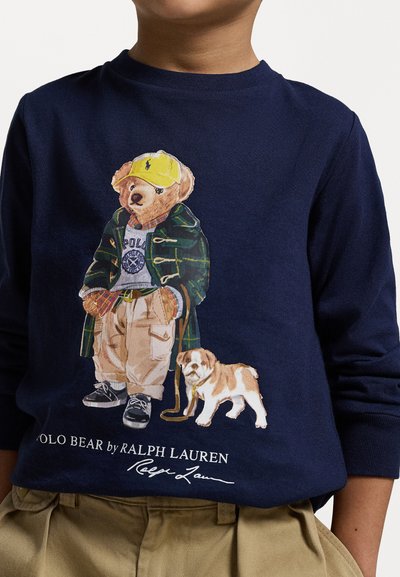 Navyblaues Langarmhemd mit einem gedruckten Grafikmotiv eines Bären in einem grünen Mantel, gelber Mütze, beigen Hosen und einem Hund neben ihm.