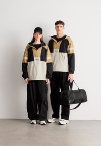 Δύο μοντέλα φοράνε μπουφάν με σχεδιασμό χρωματικών μπλοκ σε μπεζ, μαύρο και χρυσό, συνδυασμένα με μαύρο παντελόνι cargo και παπούτσια, το ένα κρατώντας μια μαύρη τσάντα.