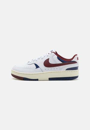 Witte lage sneaker met bordeauxrode Nike-swoosh, bordeauxrode en marineblauwe accenten, crèmekleurige zool en witte veters, gezien vanaf de buitenkant.