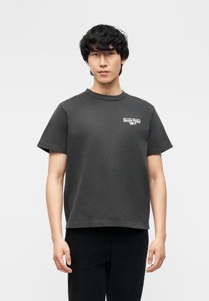 Jaunas vyras trumpais juodais plaukais, dėvintis tamsiai pilką trumpomis rankovėmis marškinėlį su mažu baltu „Service Works“ logotipu ant krūtinės.