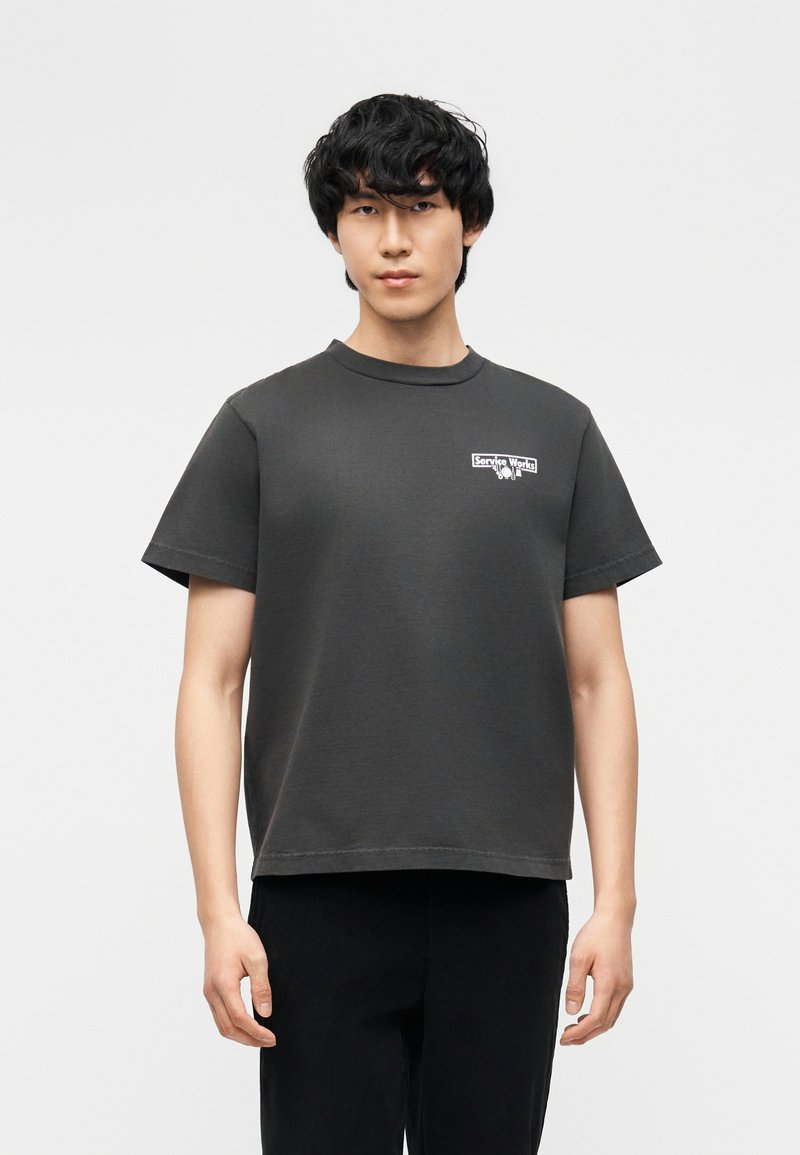 Νεαρός άντρας με κοντά μαύρα μαλλιά φορούσε σκούρο γκρι κοντομάνικο μπλουζάκι με μικρό λευκό λογότυπο "Service Works" στο στήθος.