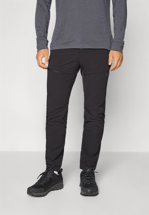 Pantalons d'extérieur noirs avec poches zippées, fabriqués à partir d'un tissu léger et flexible. Associés à des chaussures de randonnée noires. Haut gris à manches longues.