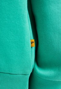 Felpa turchese con una texture liscia, polsini a coste e un piccolo etichetta gialla con un logo vicino alla cucitura.