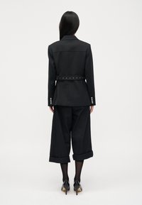 Musta räätälöity bleiseri, jossa on vyötärölle sidottu vyö, yhdistetty leveälahkeisiin mustiin housuihin. Sisältää kääntölahkeet ja tyylikkäät mustat korkokengät.