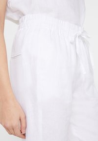 Pantalons en lin blancs dotés d'une taille élastique avec cordon de serrage, poches latérales et une texture douce, présentant une coupe décontractée dans un design minimaliste.