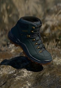 Dunkelgrüner Wanderschuh mit brauner Sohle und goldenen Ösen, auf einer felsigen Oberfläche im Freien platziert.