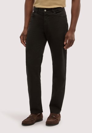 Mann trägt schwarze gerade geschnittene Hosen, beiges strukturiertes kurzärmeliges Oberteil und braune Wildleder-Loafers, steht vor einem hellen Hintergrund.