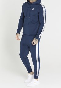 Man wearing navy blue tracksuit with white stripes down sleeves and legs, white sneakers, and visible tattoos on hands and neck.