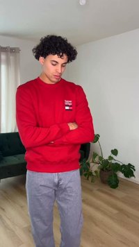 Jeune homme en pull rouge, les bras croisés, regardant vers le bas dans une pièce lumineuse avec des plantes et un canapé vert foncé.