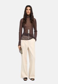 Brązowa przezroczysta bluzka z długim rękawem, luźne beżowe szerokie spodnie, czarne płaskie sandały oraz srebrny naszyjnik z zawieszką. W dłoni kopertówka.