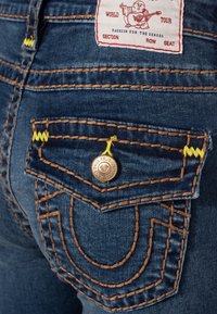 Jeans i denim har en mörkblå färg med tydlig sömnad i orange och gult. Den bakre fickan har en metallknapp och en unik design.