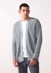 Grå stickad cardigan med öppen front, långa ärmar, två framfickor och texturerat mönster. Bärs över en vit t-shirt och grå byxor.