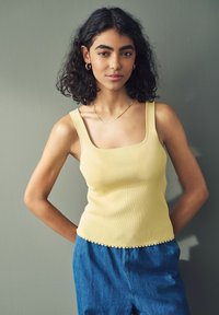 Top giallo a costine con scollatura quadrata e orlo scallop, abbinato a pantaloni in denim blu. Sullo sfondo c'è un grigio tenue.