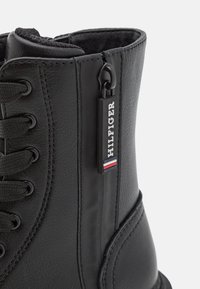Stivale in pelle nera con un design robusto, lacci frontali e un'etichetta con accenti rosso, bianco e blu. Dettagli della superficie texture visibili.