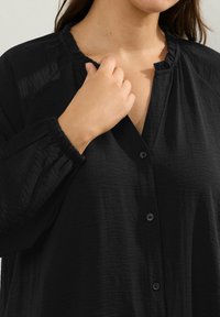 Blusa nera con bottoni, realizzata in un tessuto testurizzato, con scollatura arricciata e polsini elasticizzati. I bottoni anteriori sono in vista, mostrando una vestibilità morbida.