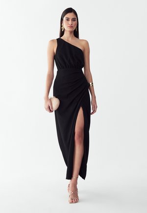 Frau trägt ein schwarzfarbenes One-Shoulder-Kleid mit hohem Schlitz, goldene Ohrringe, Armband, Riemchenabsätze und hält eine beige Clutch vor weißem Hintergrund.