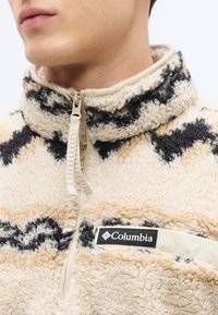 Pull polaire crème et beige avec motif zigzag noir, col montant, fermeture éclair sur le devant et patch logo sur la poche poitrine.