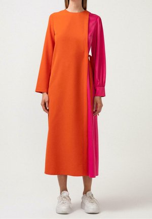 Femme portant une robe à manches longues couleur bloc avec devant et manches orange, et un panneau latéral et manches rose vif, assortie à des baskets blanches.