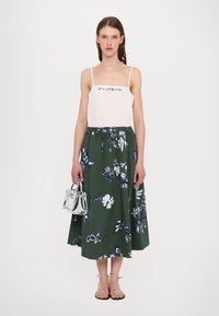Top alb cu bretele subțiri, decorat cu broderie florală, asortat cu o fustă lungă verde cu modele florale mari. Ține o geantă argintie.